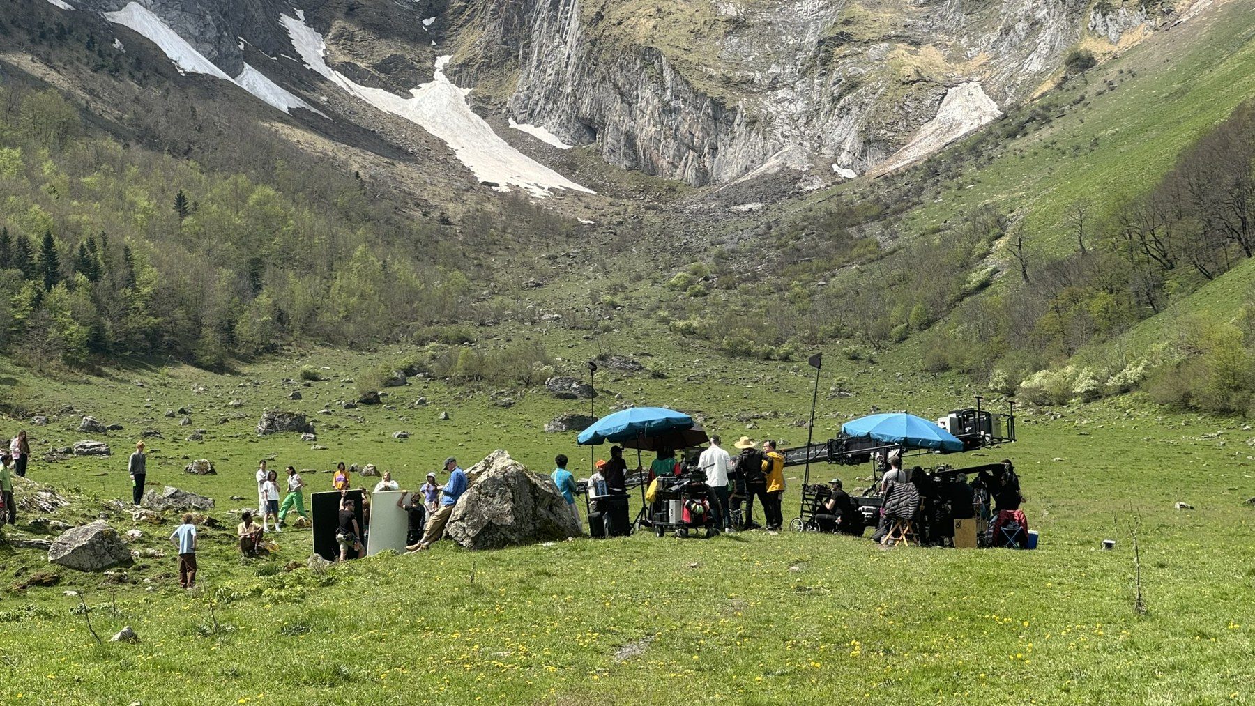 Equipo de rodaje con grúa, monitores y sombrillas en un prado bajo montañas nevadas