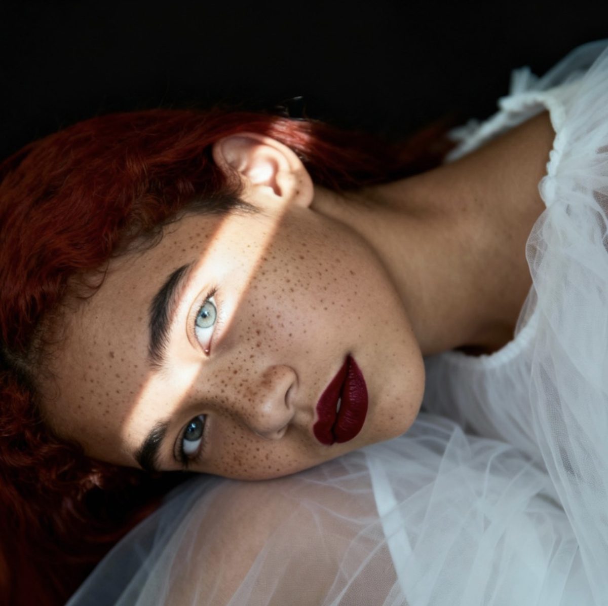Retrato de una chica pelirroja con pecas y labios granate tumbada sobre tul blanco, con una franja de luz en los ojos